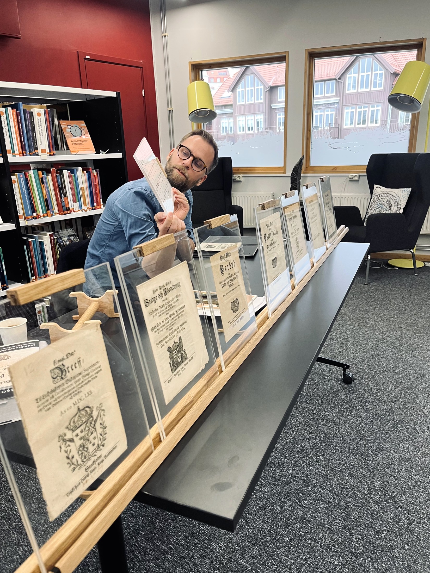 En person bakom ett bord med historiska originaltryck uppställda i träställ, med bokhyllor i bakgrunden.