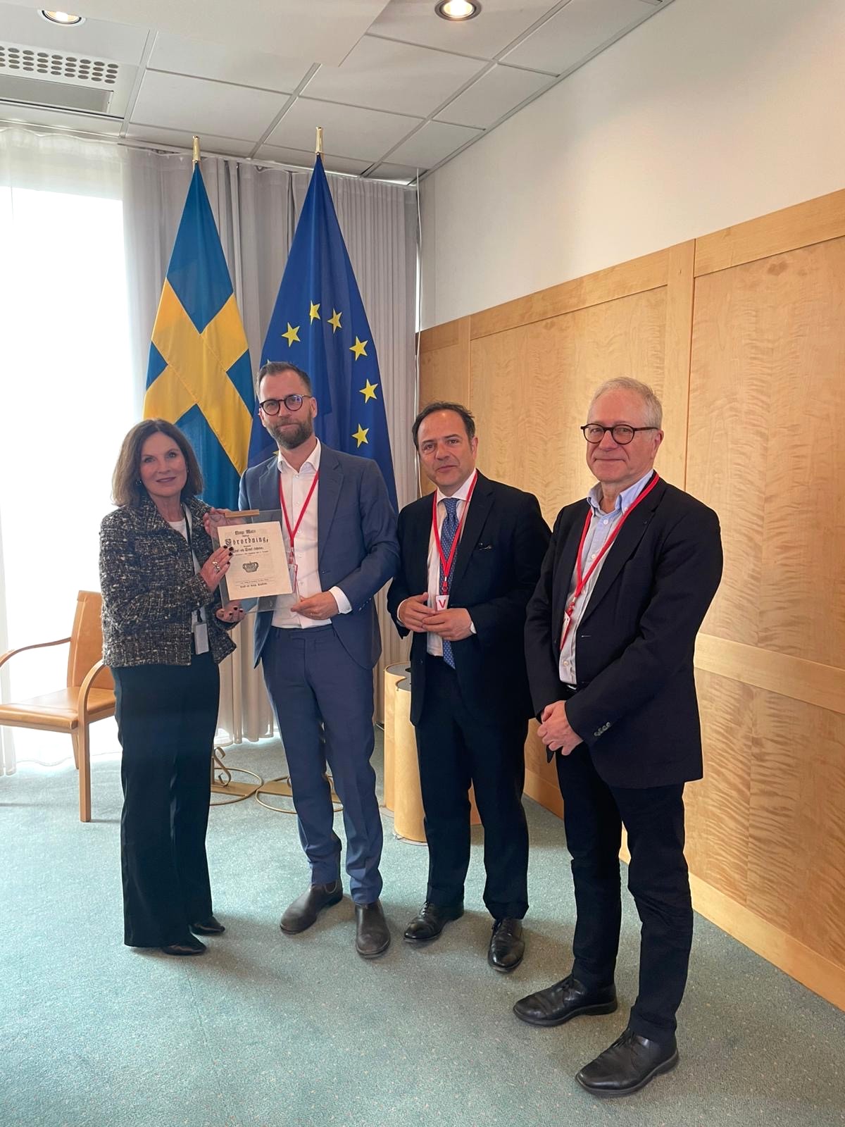 Fyra personer står tillsammans med ett inramat dokument framför svenska och europeiska flaggor.